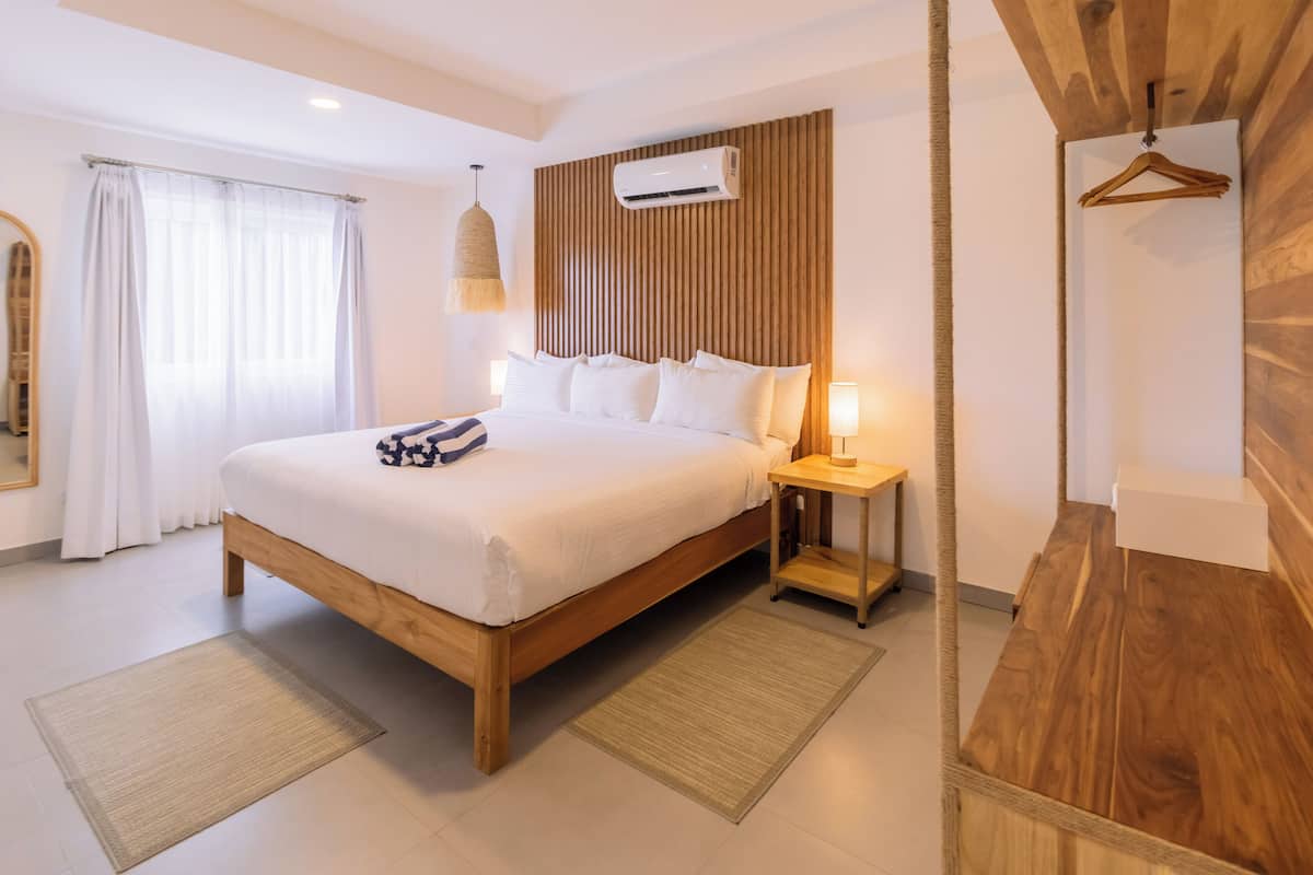 A modern hotel room with a wooden bed, a bedside table with a lamp, a hanging light fixture, and a wooden panel wall.