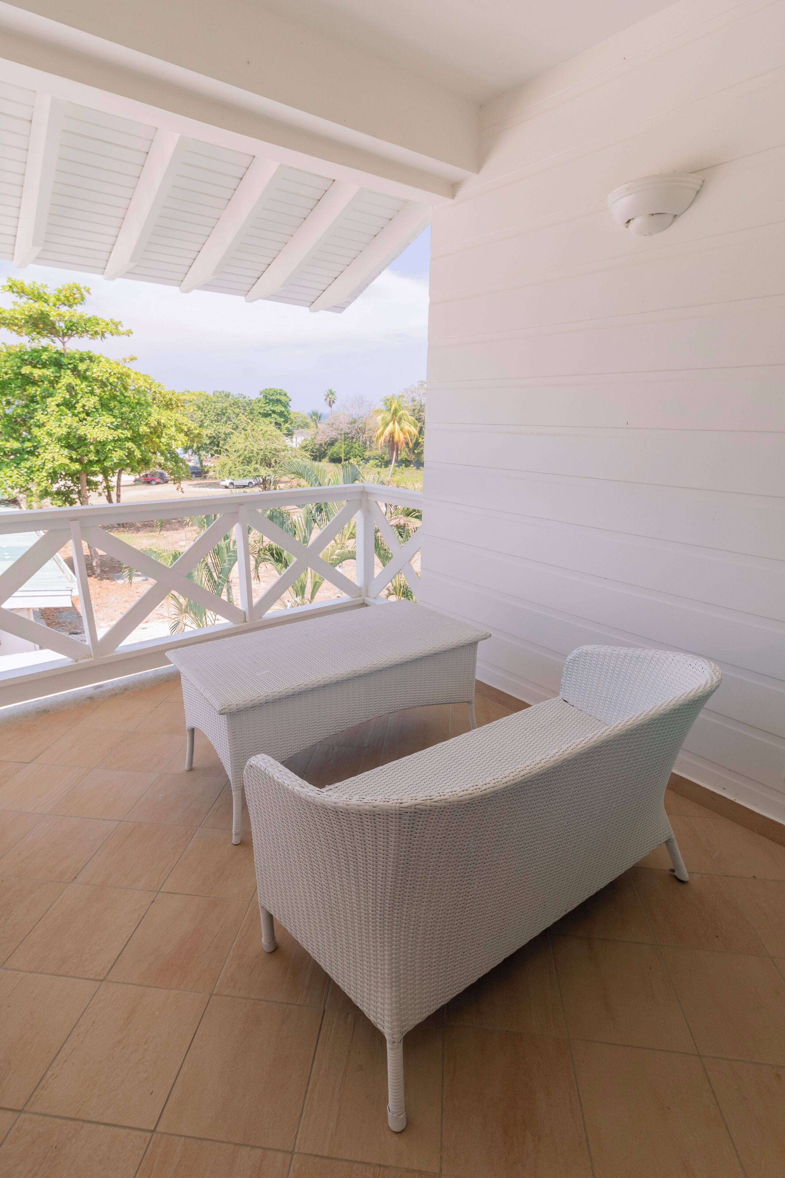 A covered patio with wicker furniture, a white railing, and a view of greenery and palm trees.