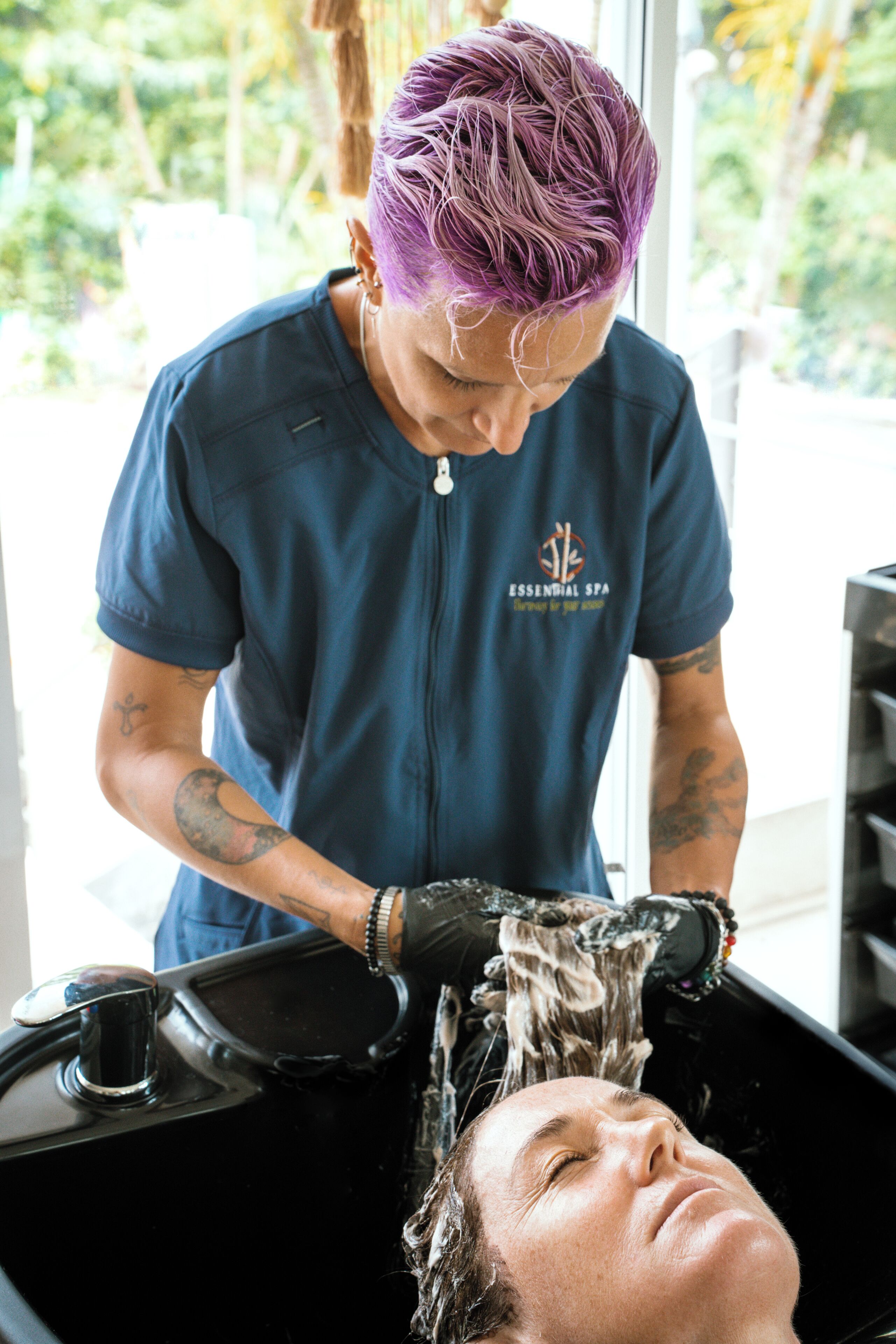 A person with purple hair washing another person's hair in a salon.