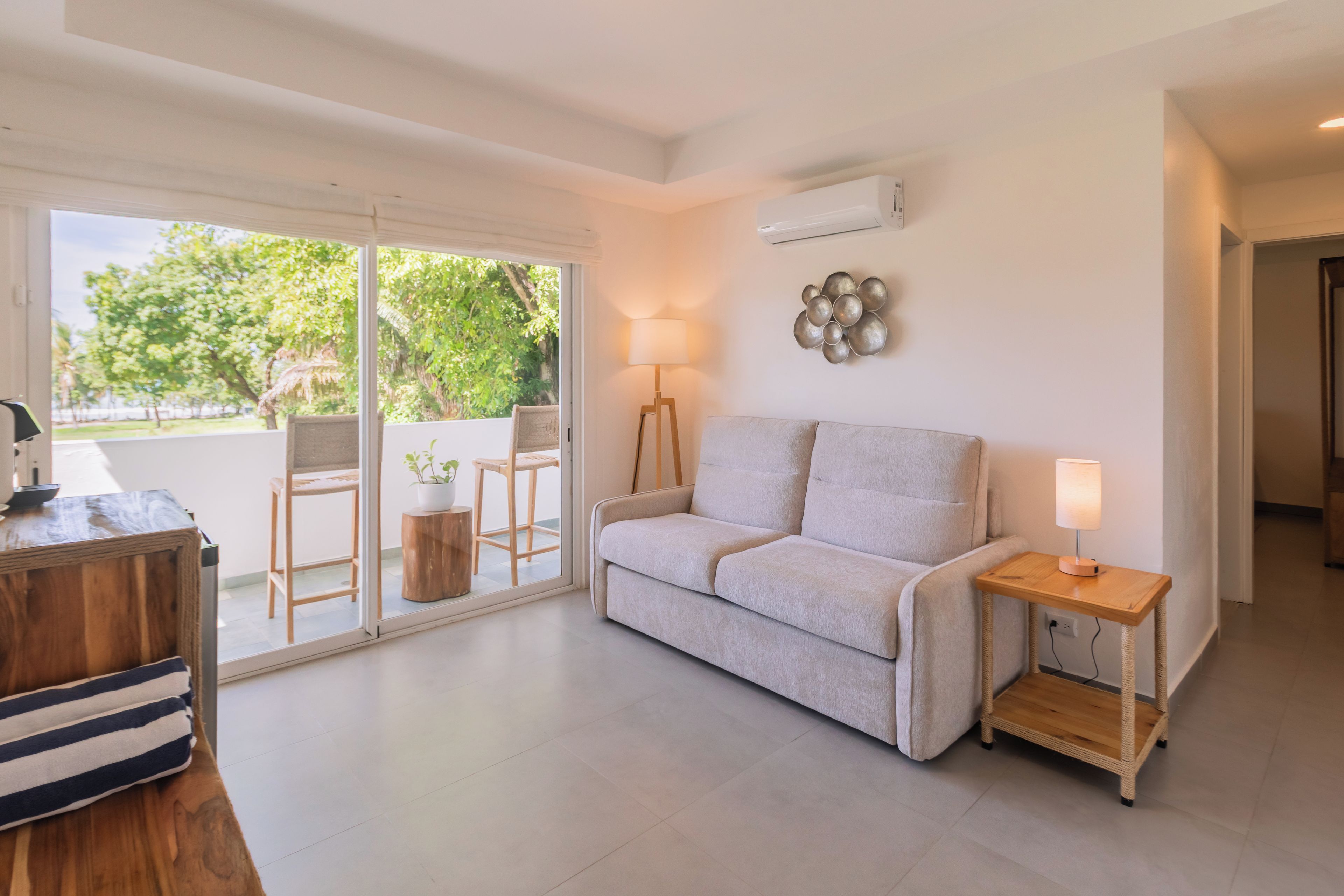 A modern living room with a sofa, a small wooden table, a lamp, and a view through a glass door to an outdoor area with chairs and greenery.
