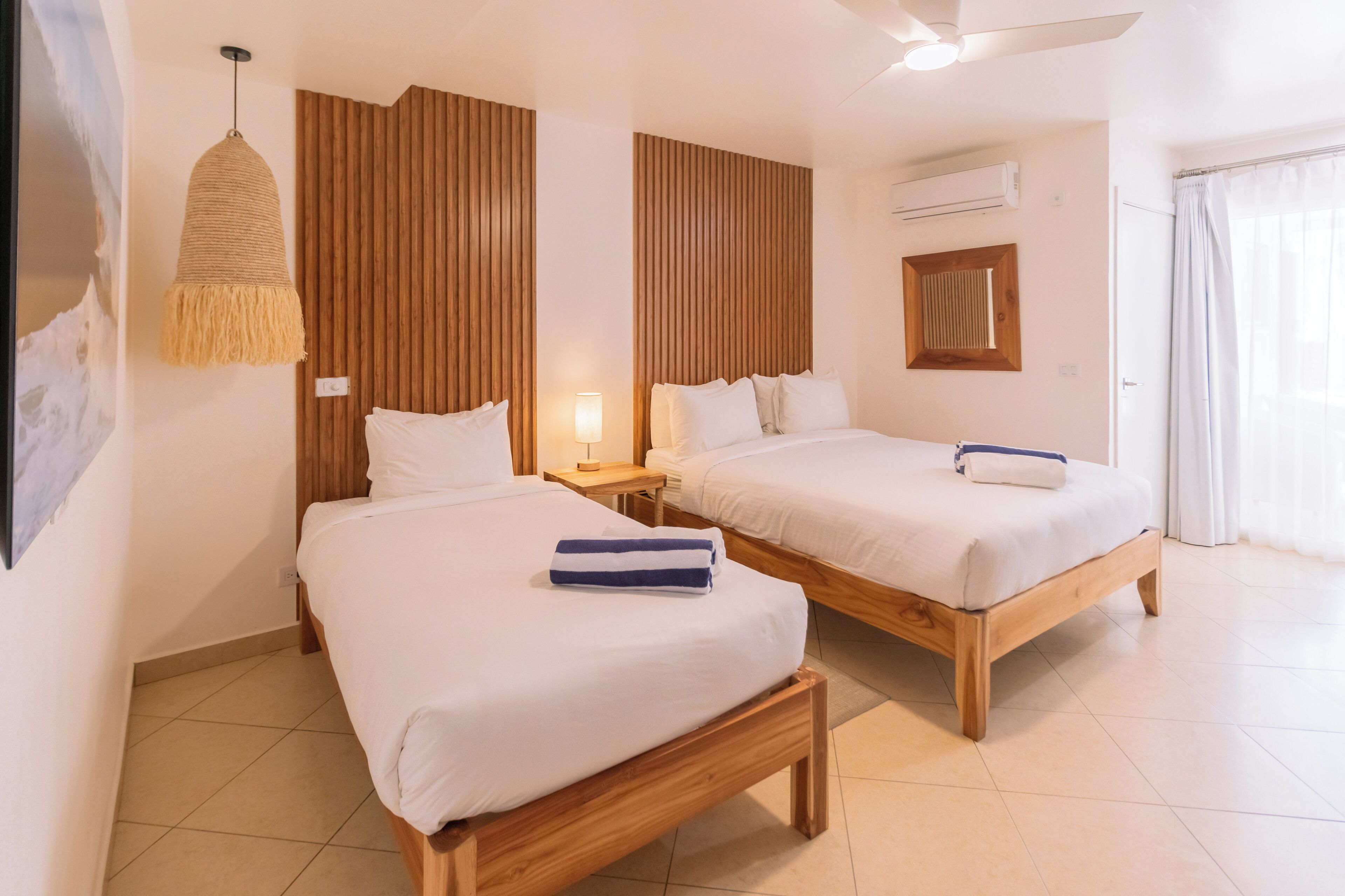 A hotel room with two wooden beds, a hanging light fixture, a wall-mounted air conditioner, and a window with curtains.