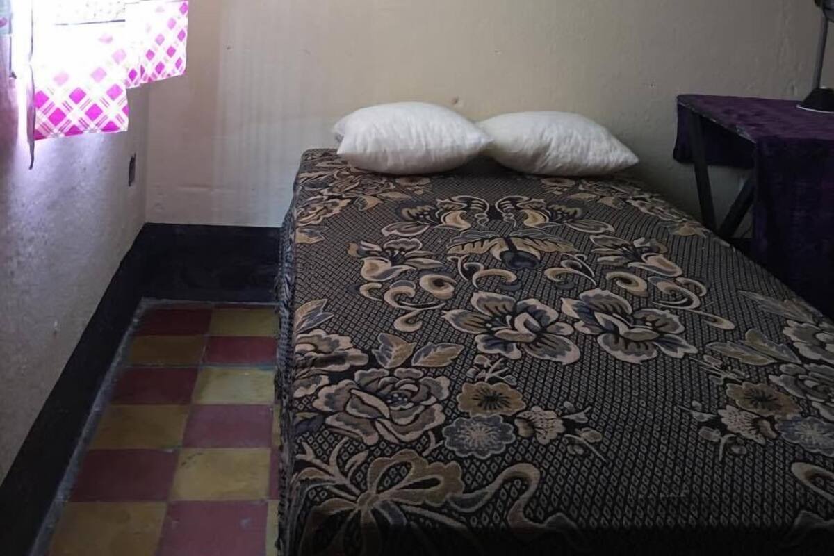 Une petite pièce simple, avec un lit, une table et une fenêtre ornée d’un rideau à motifs floraux.
