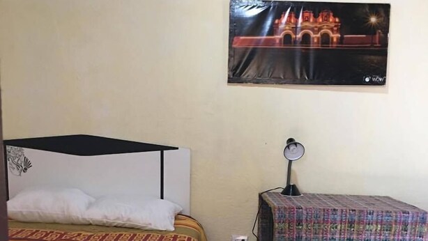 Een kamer met een bed, nachtkastje, lamp, een tafelkleed met dessin en een aan de muur bevestigde televisie waarop een gebouw te zien is.