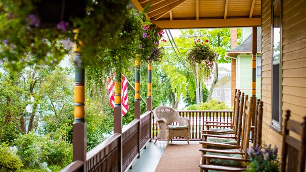 A covered porch with wicker chairs and a hanging chair, surrounded by greenery and a view of a house.