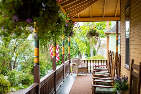 A covered porch with wicker chairs and a hanging chair, surrounded by greenery and a view of a house.