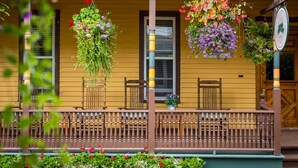 A yellow building with a porch, wooden chairs, and hanging flower baskets.