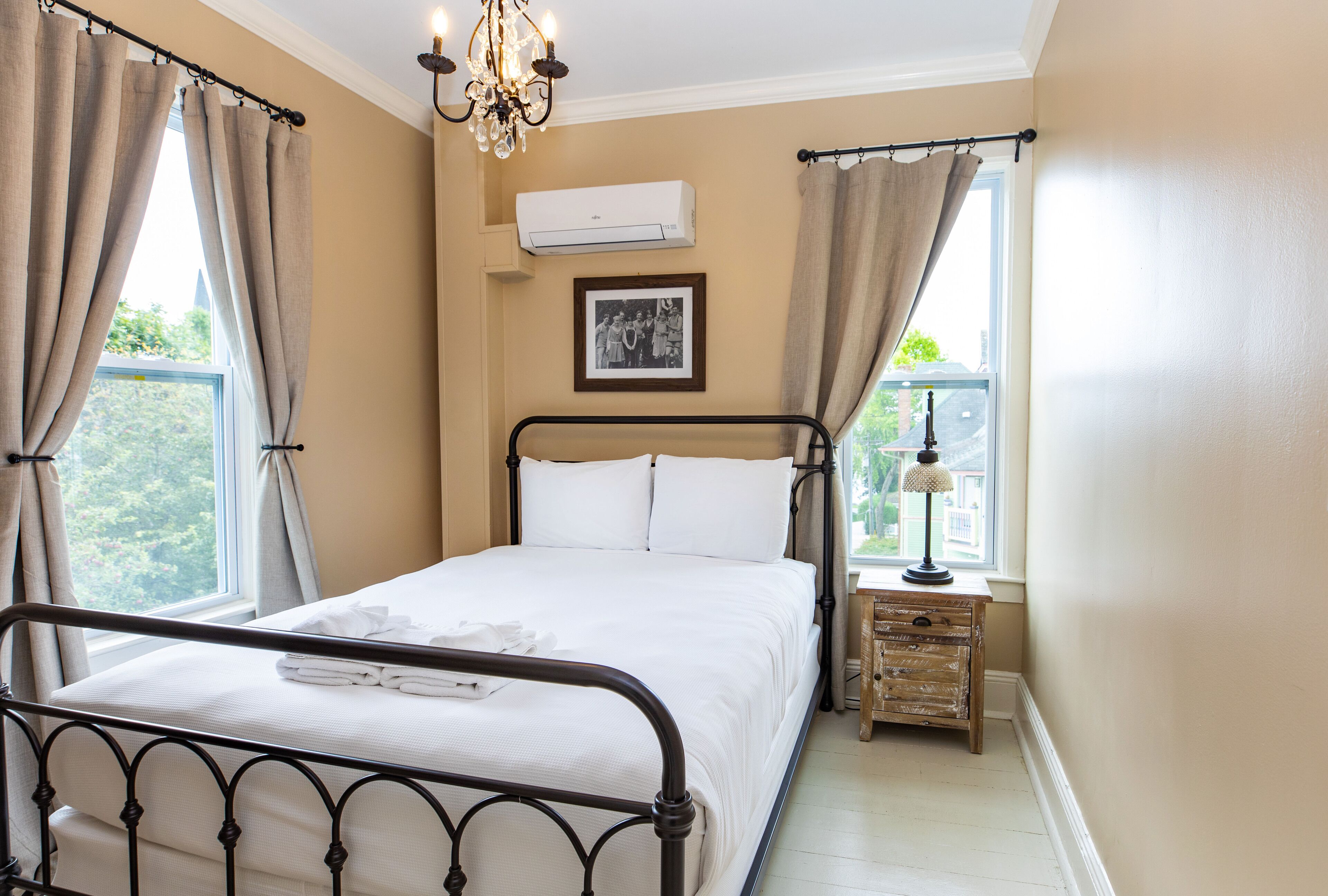 Un dormitorio con cama, cortinas, aire acondicionado, mesita de noche con lámpara y un cuadro en la pared.