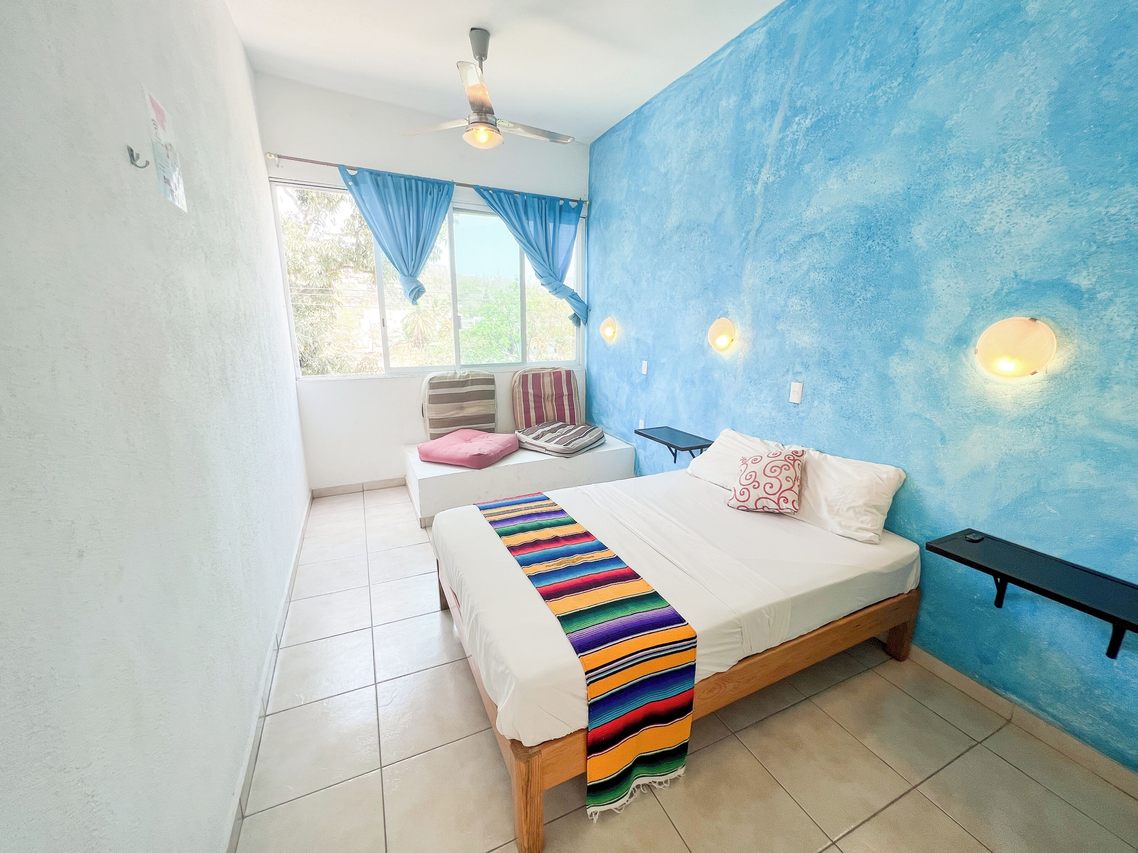 A bedroom with a bed, a bench, a window with curtains, a ceiling fan, and wall-mounted lights.