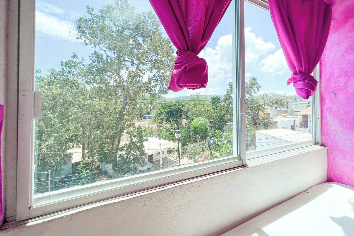 A window with pink curtains, a view of trees and buildings outside.