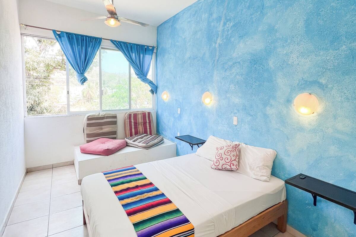 A bedroom with a bed, a ceiling fan, a window with curtains, and a wall-mounted light fixture.