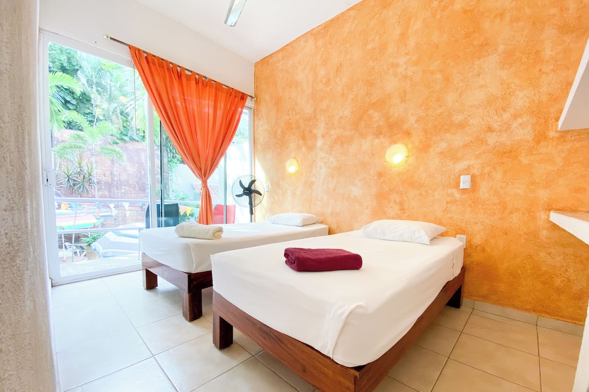 A hotel room with two beds, a sliding glass door leading to an outdoor area, and a fan on the left side.