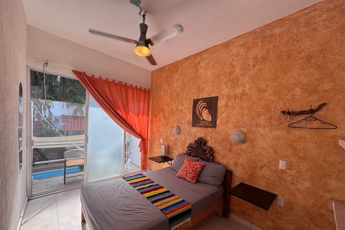 Een slaapkamer met een bed, een plafondventilator, een aan de muur bevestigde lamp, een raam met gordijnen en uitzicht op het omliggende landschap.