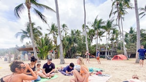 A group of people sitting on beach mats under palm trees, with a tent and beach chairs in the background.