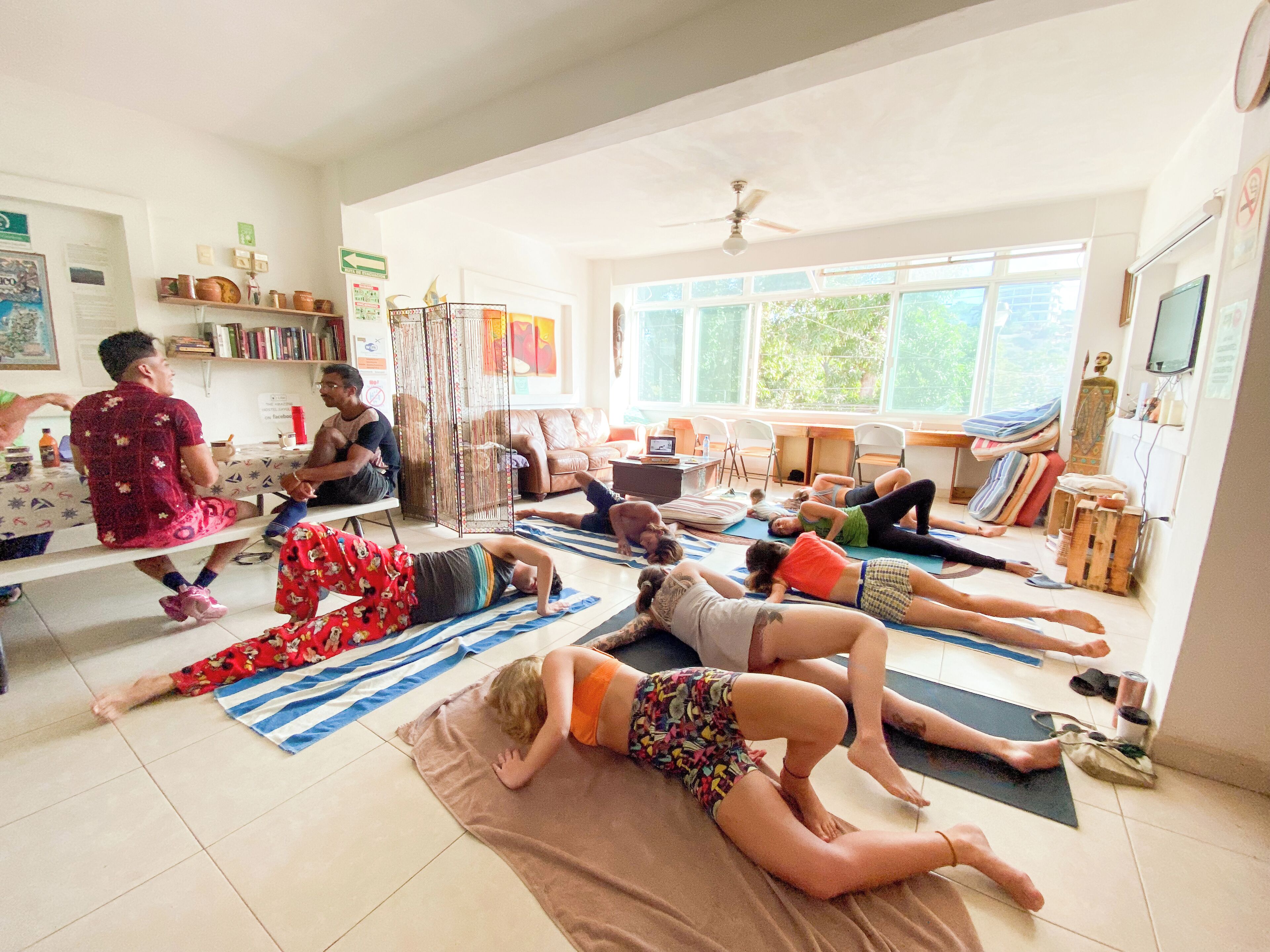 Ein Yoga-Kurs in einem hell erleuchteten Raum mit großen Fenstern, einem Deckenventilator sowie verschiedenen Yogamatten und Zubehör.