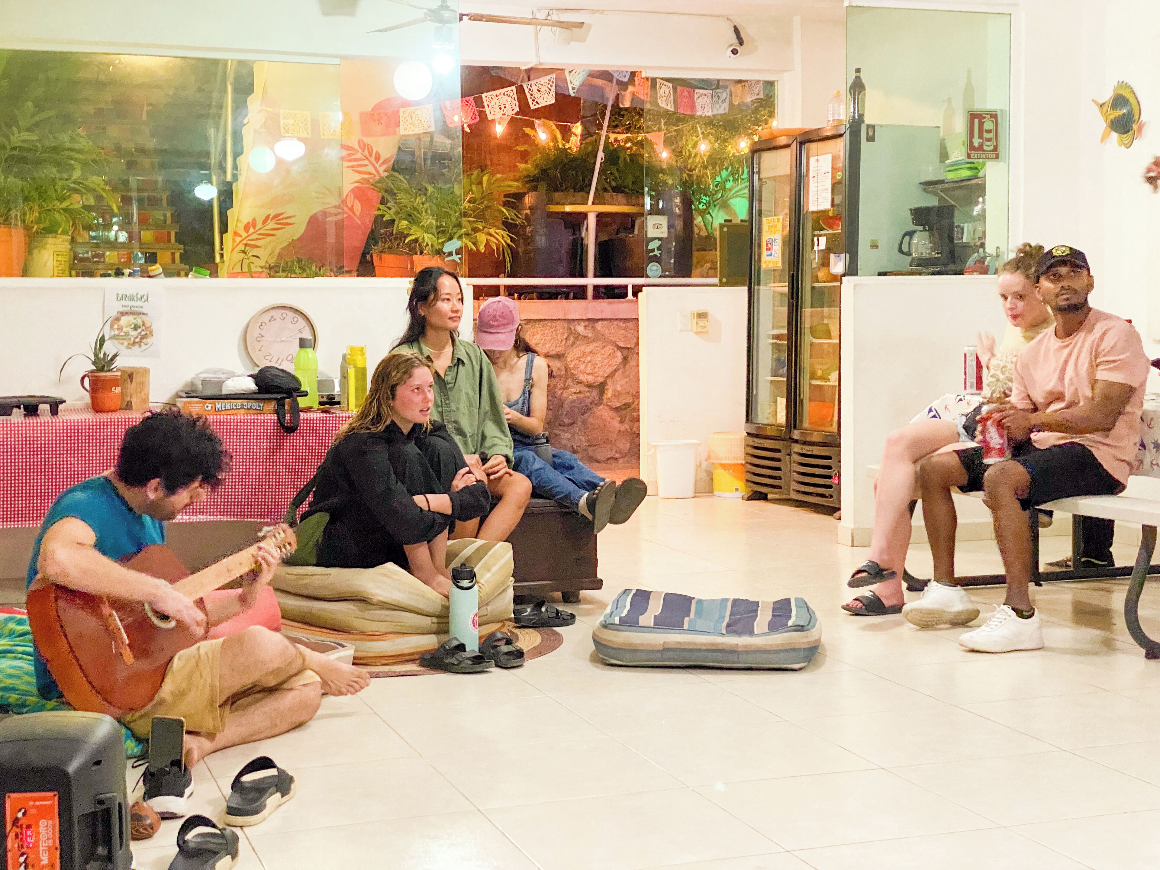 A group of people sitting on the floor in a room with a kitchen area visible through a glass door.