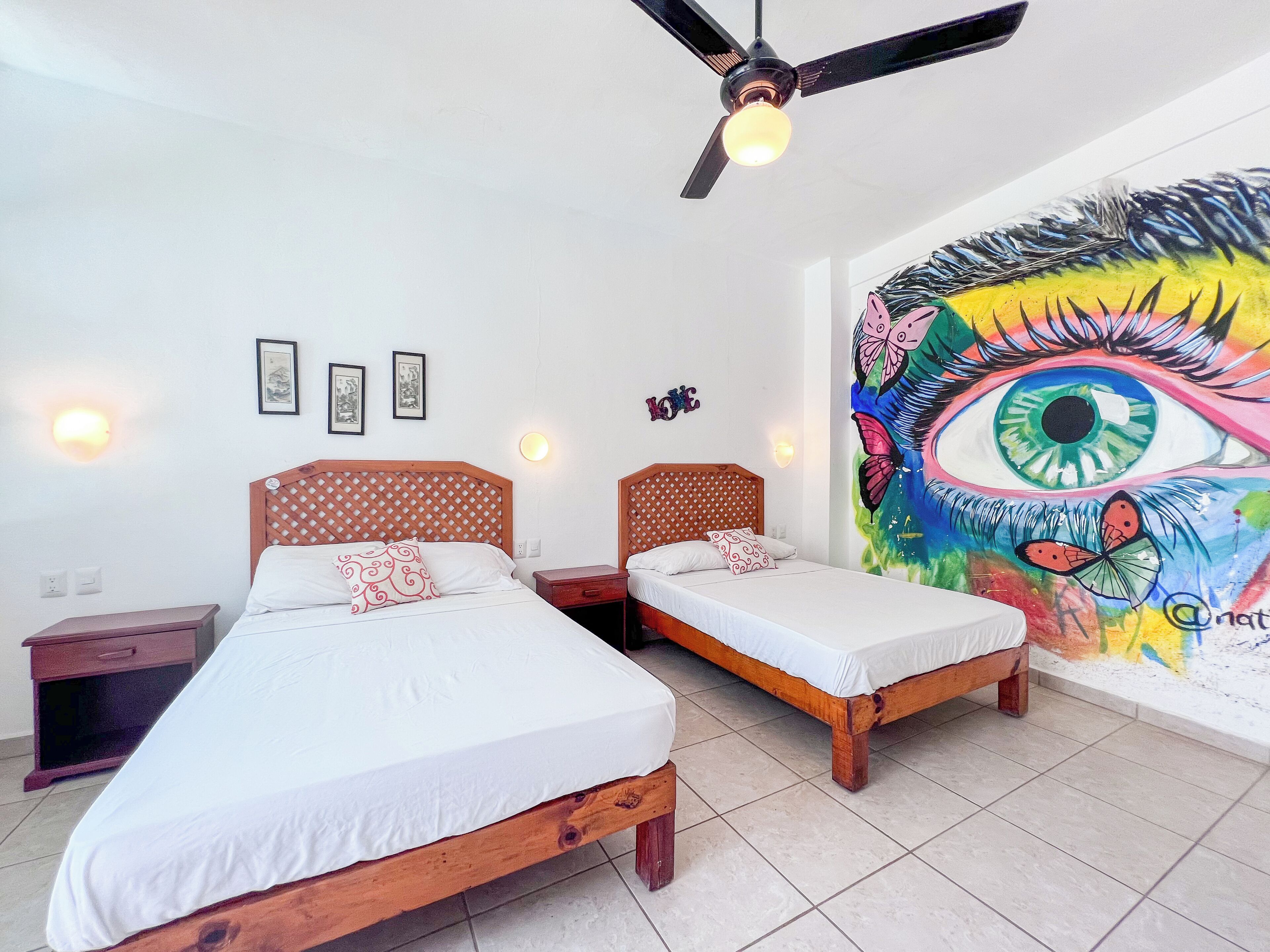 A room with two beds, a ceiling fan, wall-mounted lights, and a large colorful mural.