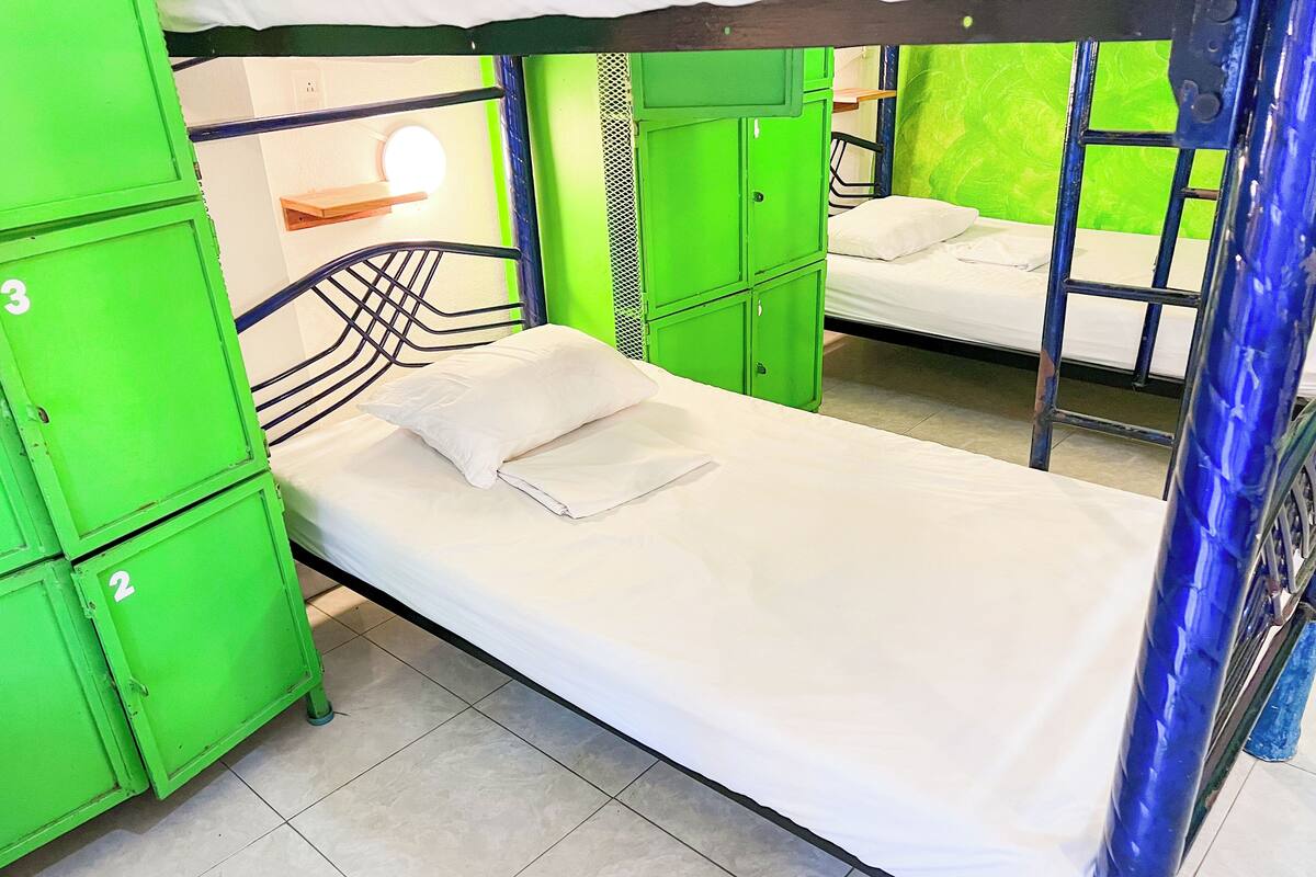 A dormitory room with bunk beds, green lockers, and a white bed with a folded blanket.