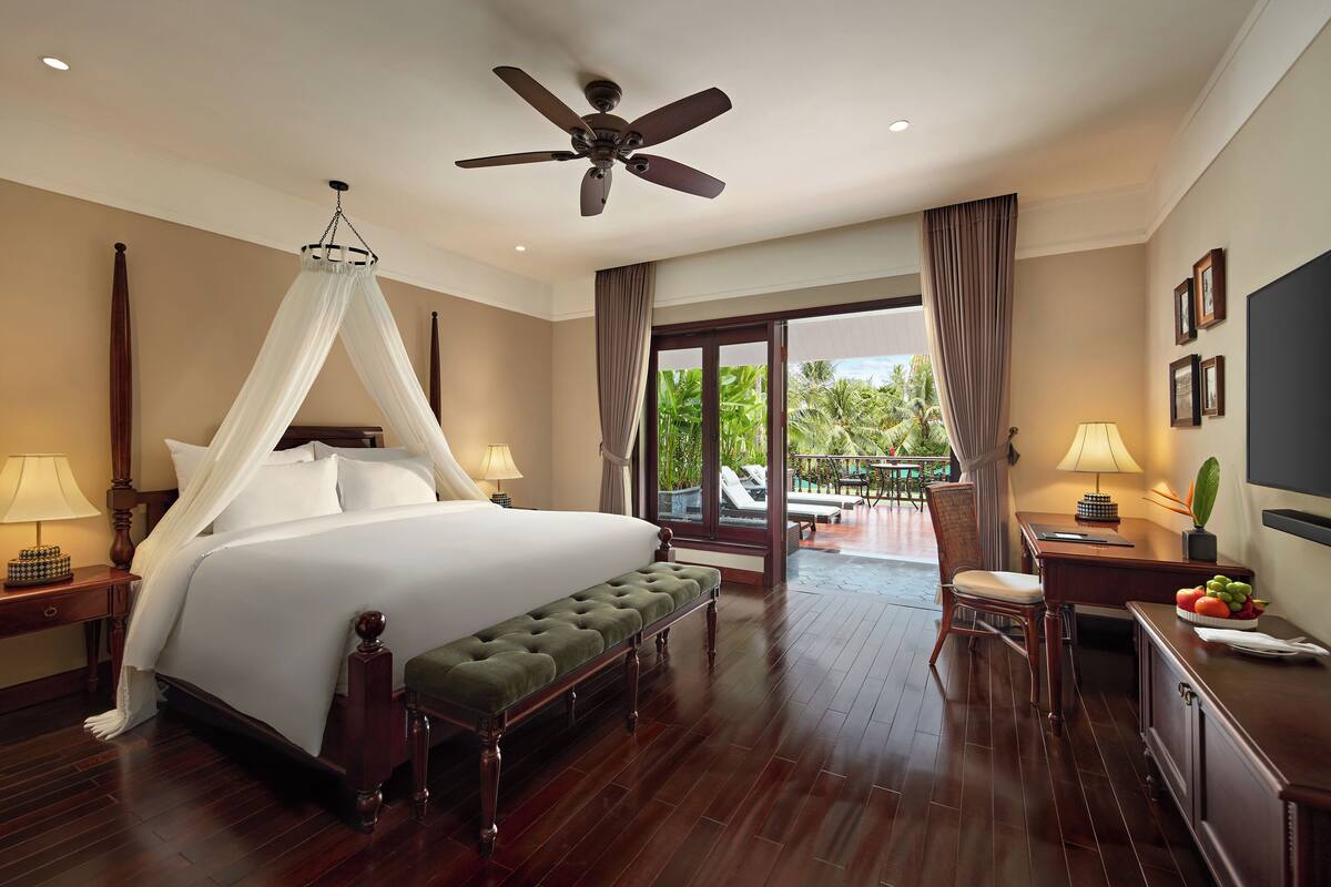 Habitación de hotel con una cama grande, ventilador de techo, un banco, escritorio con lámpara y vistas al exterior.