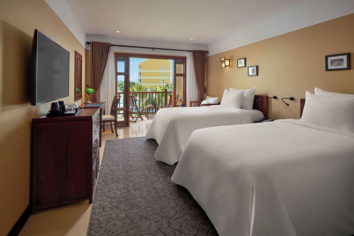 Habitación de hotel con dos camas, televisión, escritorio con lámpara y balcón con vista a edificios y follaje.