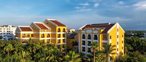 A yellow building with multiple balconies and a red roof, surrounded by palm trees and other greenery.
