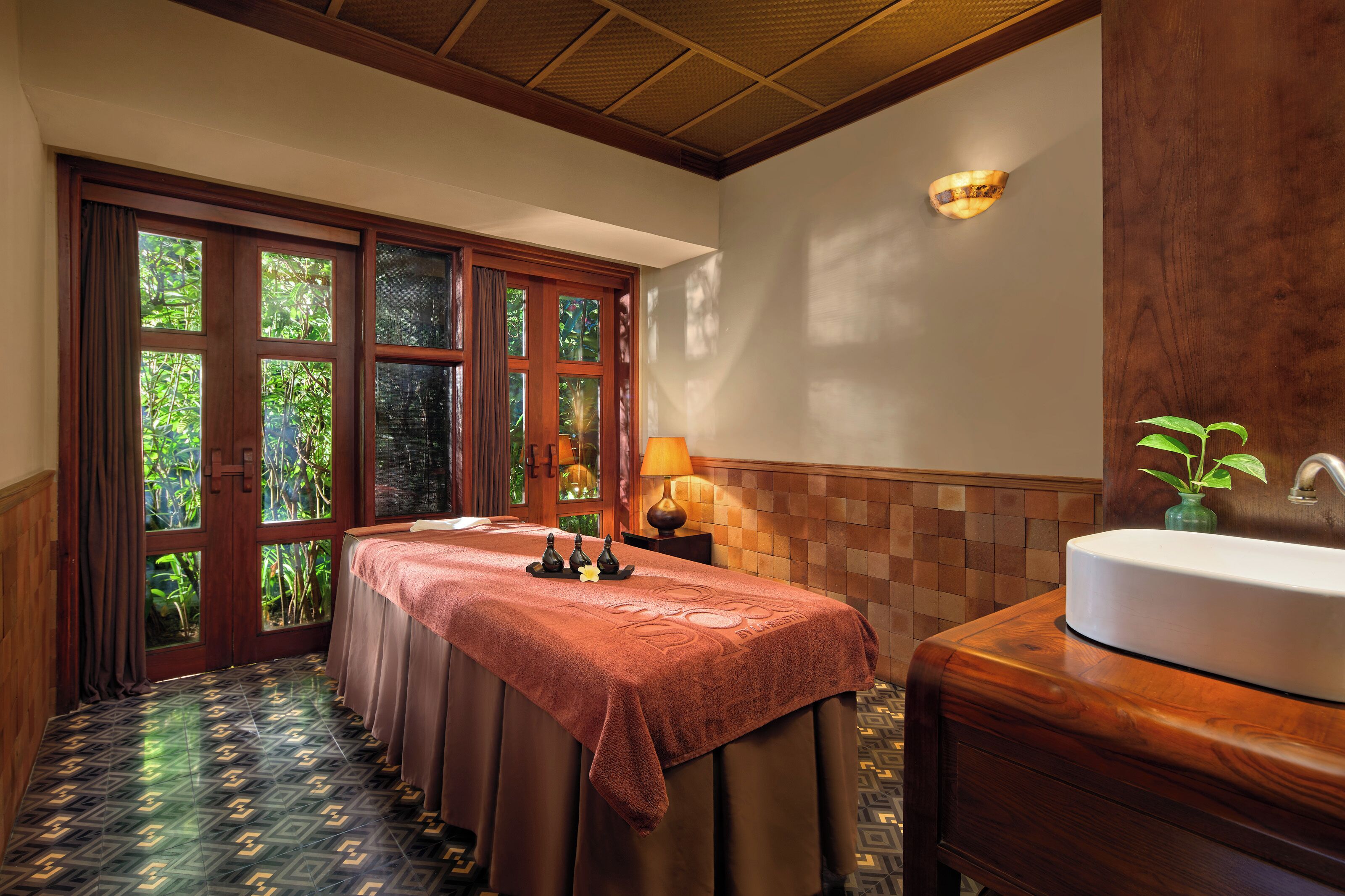 A room with a massage table, a sink, and a view of greenery through the windows.