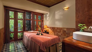 A room with a massage table, a sink, and a view of greenery through the windows.