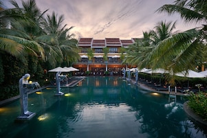 A resort pool with palm trees, a building with a tiled roof, and lounge chairs.