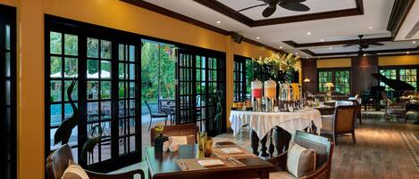 A well-lit dining area with tables set for guests, large windows offering a view of the outdoors, and ceiling fans for comfort.