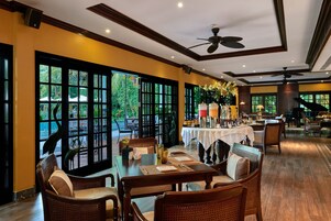 A well-lit dining area with tables set for guests, large windows offering a view of the outdoors, and ceiling fans for comfort.