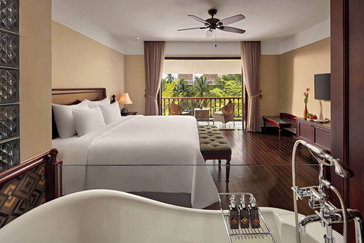 Una habitación de hotel con una cama grande, una bañera, un balcón con muebles de mimbre y un televisor en la pared.