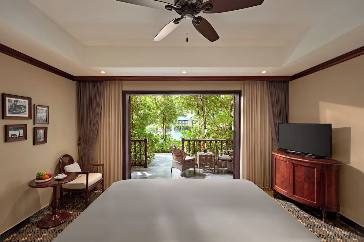 Habitación de hotel con una cama grande, un televisor sobre un mueble de madera, un área de comedor con sillas de mimbre y un balcón con vista a follaje.