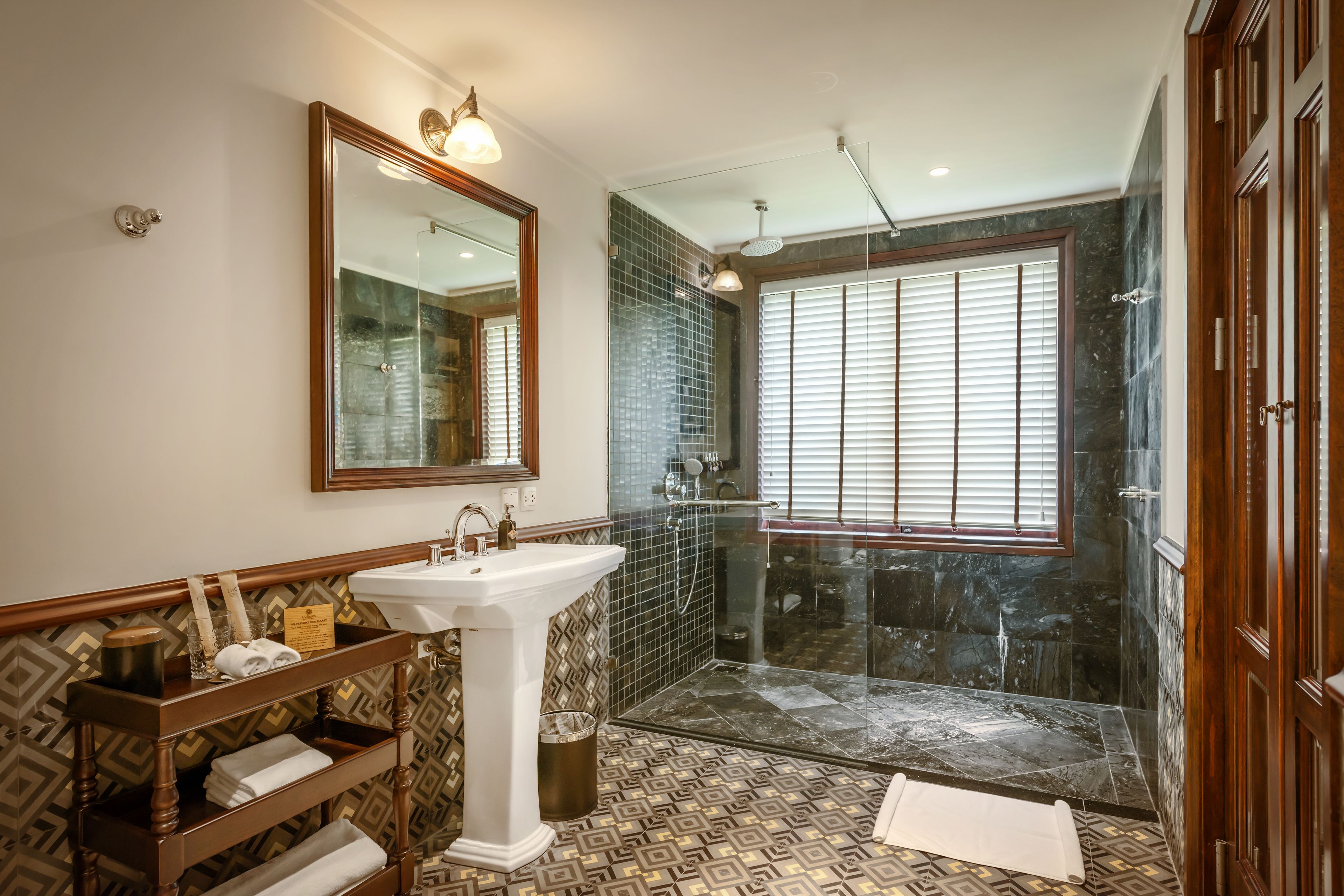 Un baño con un espejo grande, un lavabo, un mueble de tocador con toallas, una ducha con puerta de cristal y baldosas de suelo estampadas.