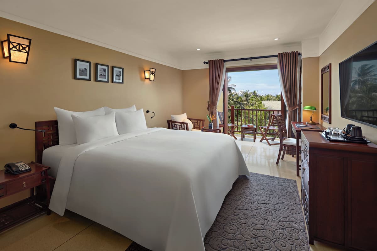Habitación de hotel con una cama grande, un escritorio con teléfono, una silla, un balcón y vistas a palmeras.