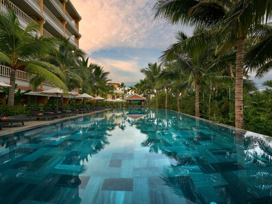 A swimming pool with lounge chairs and umbrellas, surrounded by palm trees and a hotel building.