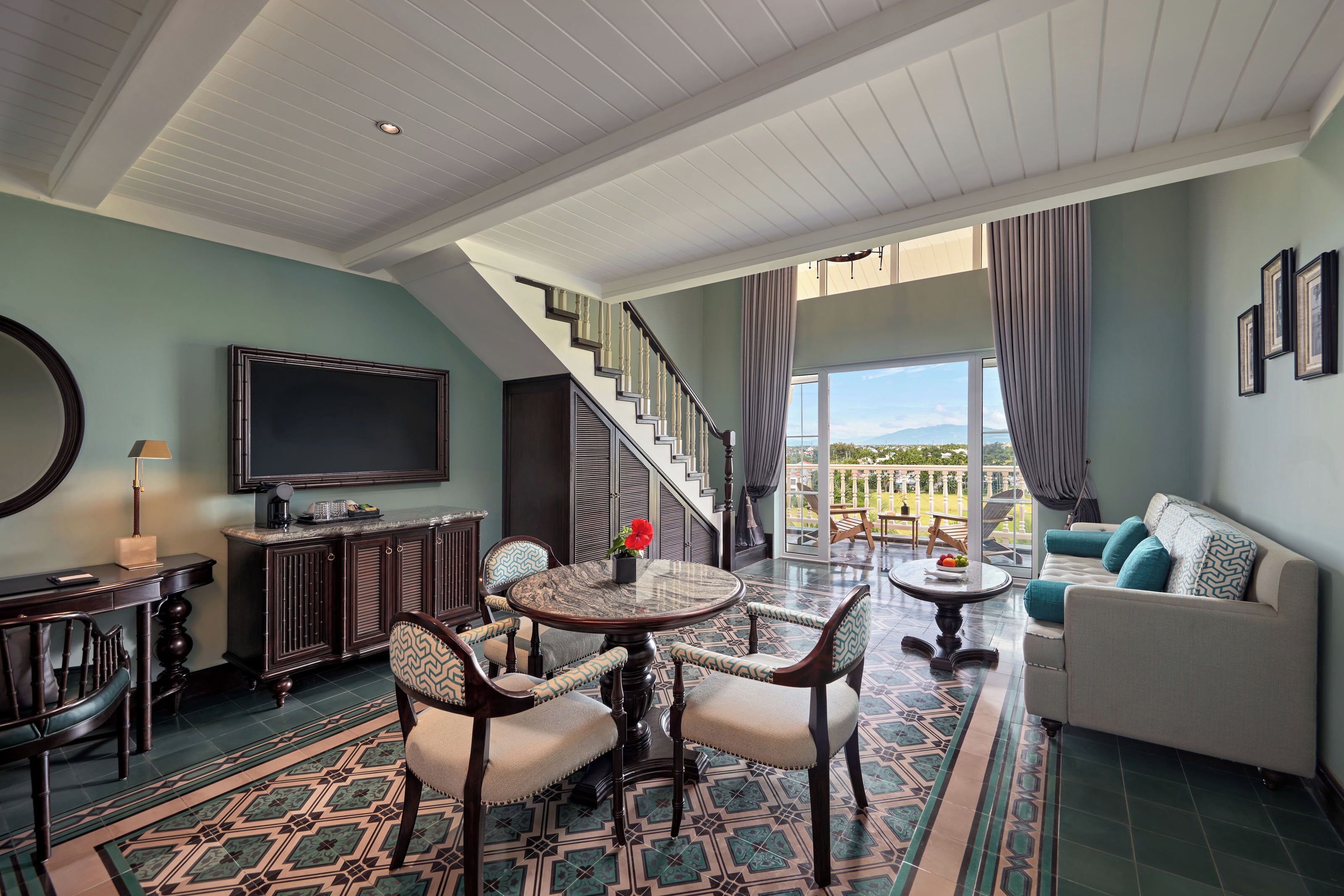 A spacious living room with a large TV, a dining area with a round table, and a staircase leading to a balcony with a view.