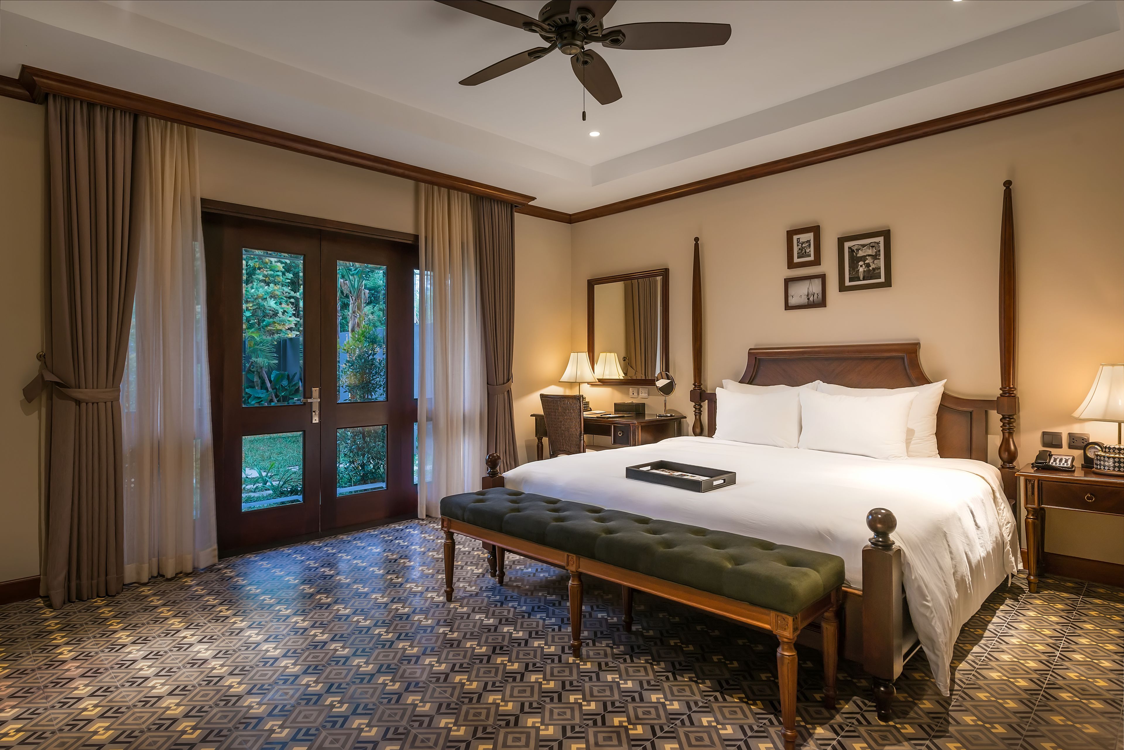A hotel room with a large bed, a bench, a ceiling fan, a nightstand, a lamp, and a door with a view of greenery.