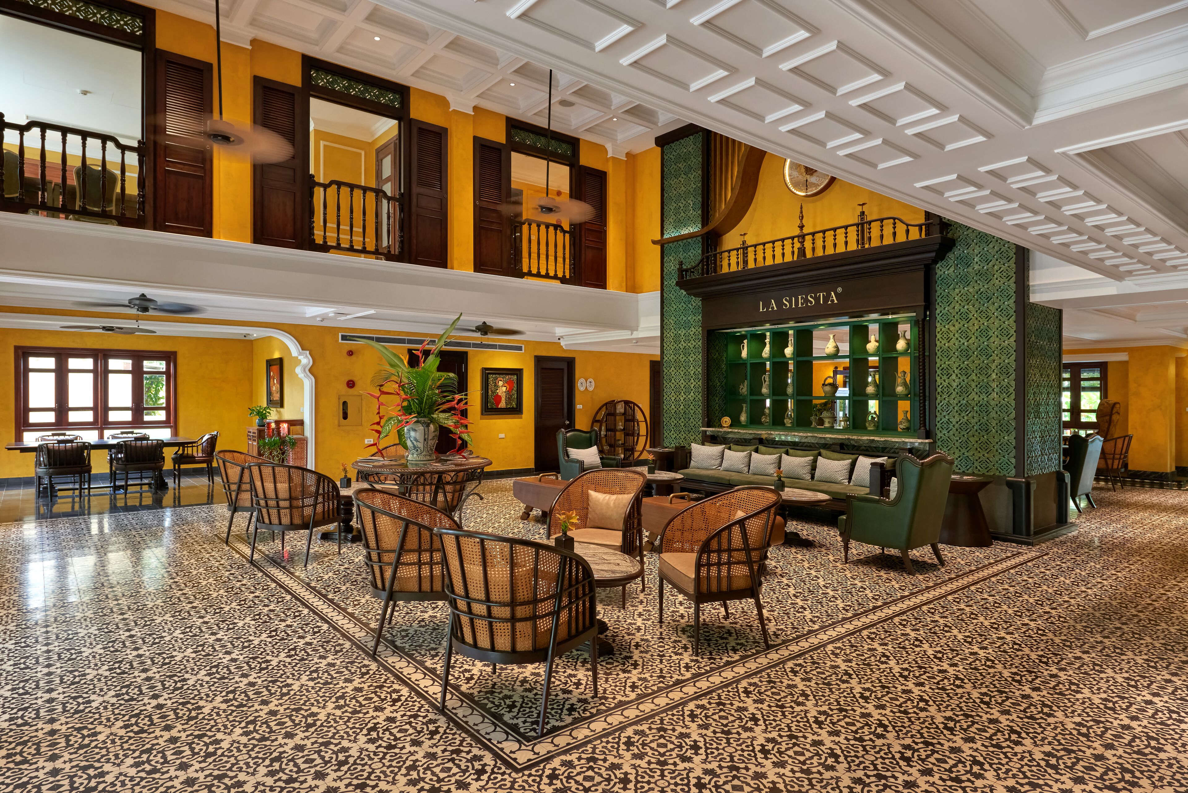 A spacious lobby with a high ceiling, a central seating area with wicker chairs, a bar named 'LA SESTA', and a patterned floor.