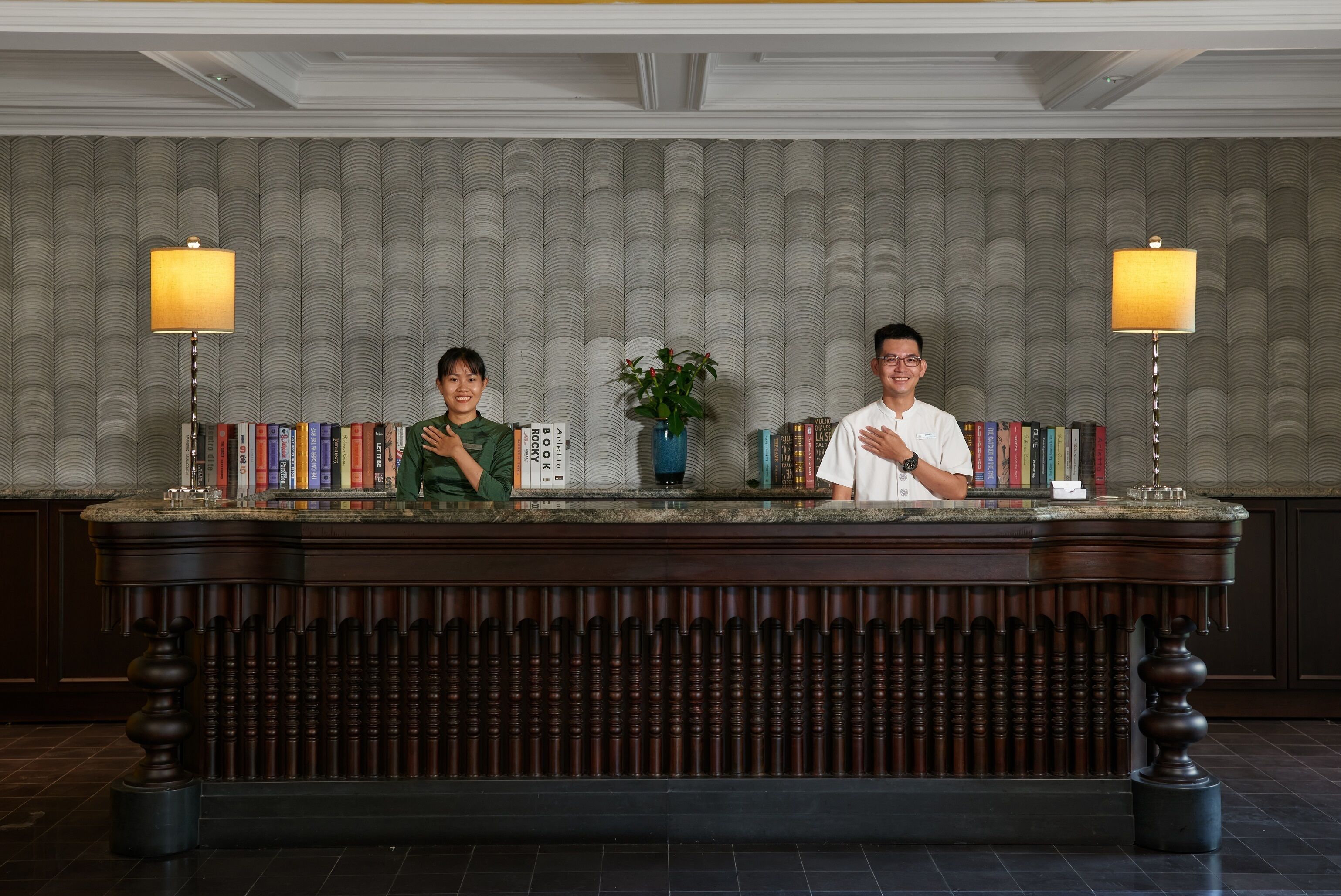 A reception desk with two individuals standing behind it, surrounded by books and a potted plant.
