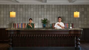 A reception desk with two individuals standing behind it, surrounded by books and a potted plant.
