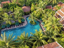 An aerial view of a resort with a central swimming pool, surrounded by palm trees and lounge areas with umbrellas.