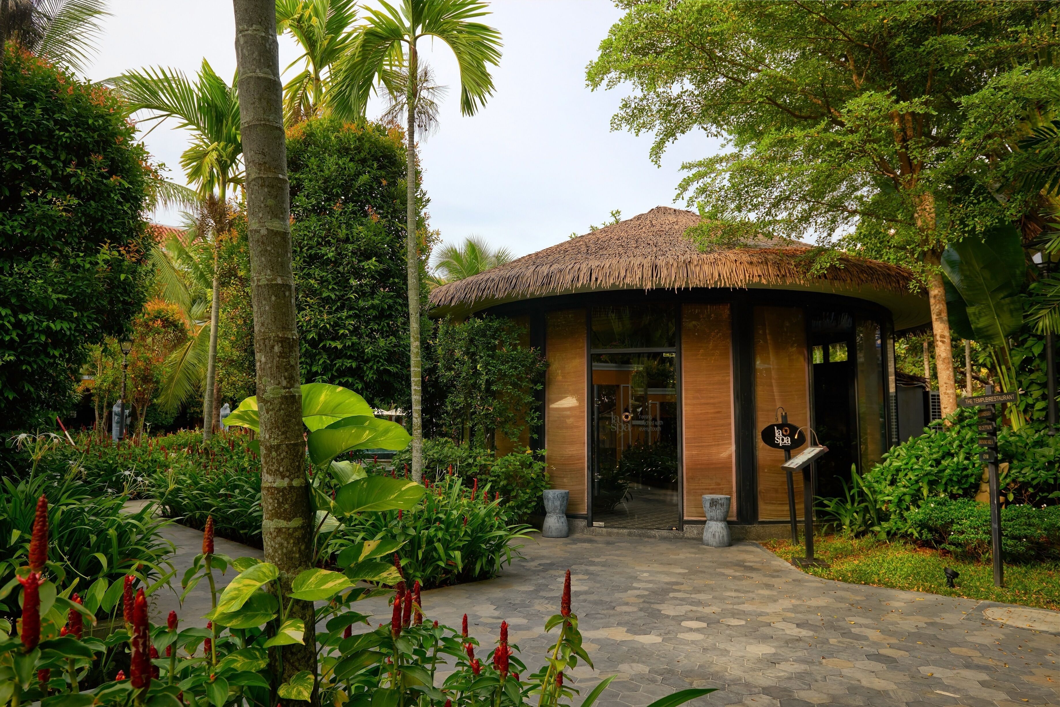 A tropical garden entrance with a thatched-roof structure, paved pathway, and lush greenery.