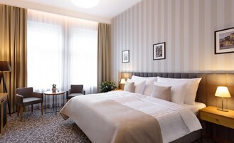 Una camera d'albergo con un letto grande, due comodini con lampade, un tavolino rotondo con una sedia e un'ampia finestra con tende.