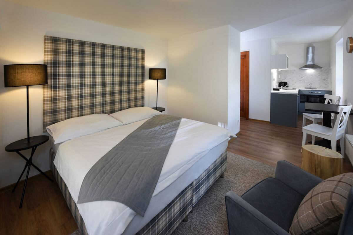 Ett modernt hotellrum med en stor säng, sänglampor, ett litet runt bord, en grå soffa och en köksdel med spishäll och diskho.