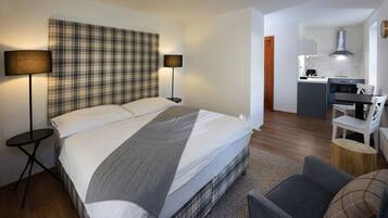 A modern hotel room with a large bed, bedside lamps, a small round table, a grey sofa, and a kitchen area with a stove and sink.