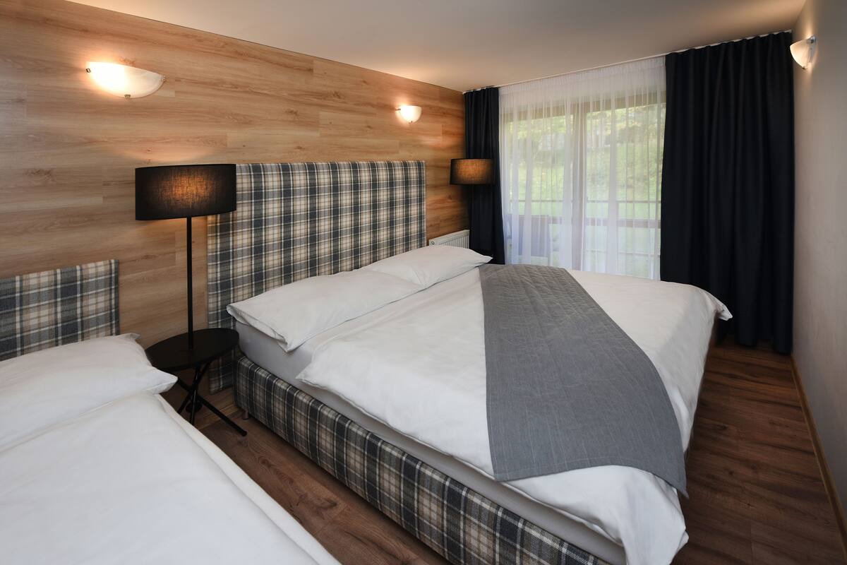 Ett modernt hotellrum med en sänggavel i trä, en stor säng med vita sängkläder, ett sängbord med en lampa och utsikt över grönskan genom tunna gardiner.