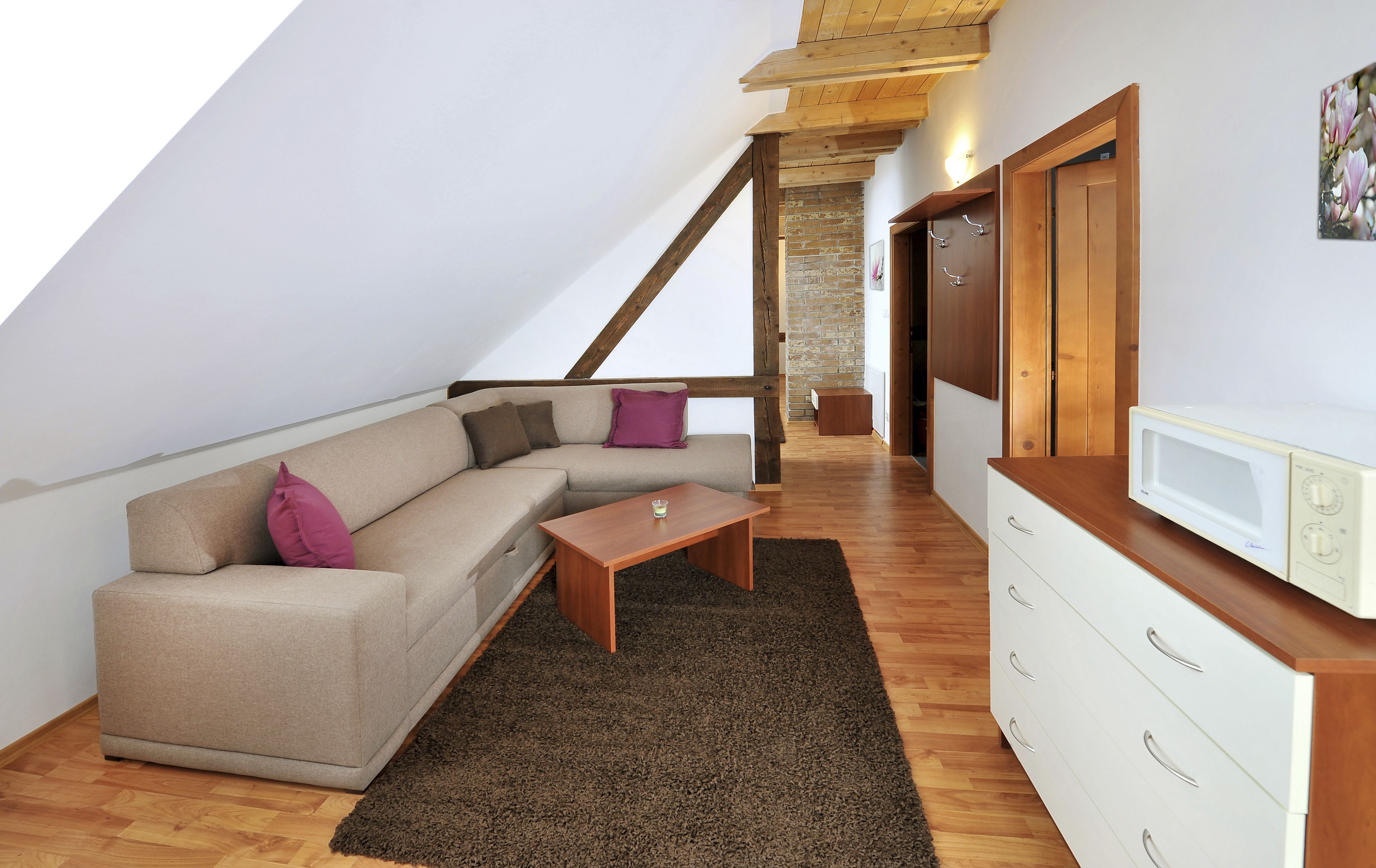 Une pièce de style loft avec un plafond en bois, un canapé d’angle beige, une petite table en bois, une commode blanche et un four à micro-ondes.