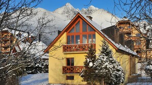 Un edificio amarillo con techo rojo y balcón de madera, rodeado de árboles nevados y montañas.