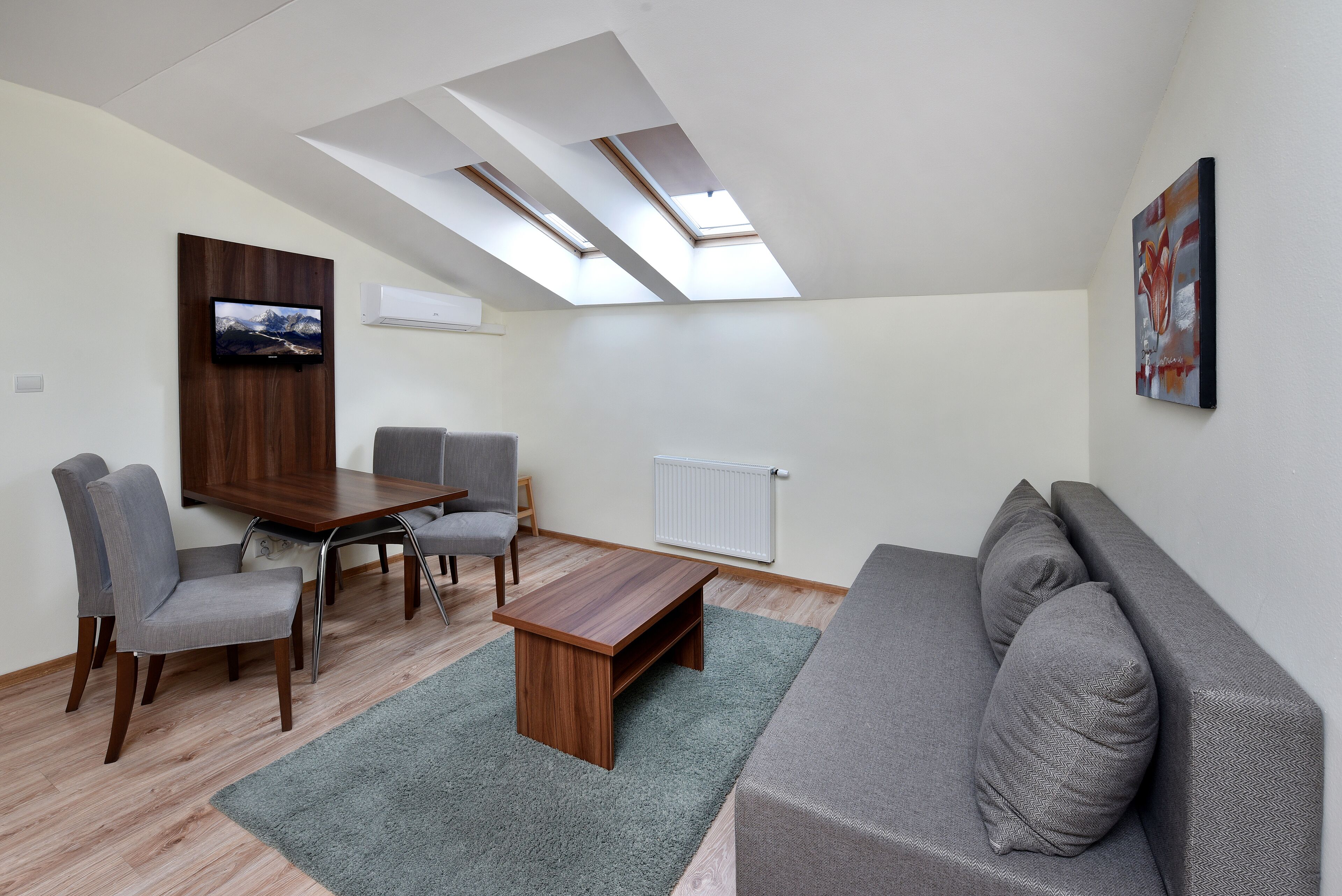 A modern living room with a grey sofa, a wooden coffee table, and a dining table with chairs. There are skylights on the ceiling, a mounted TV, and a radiator.