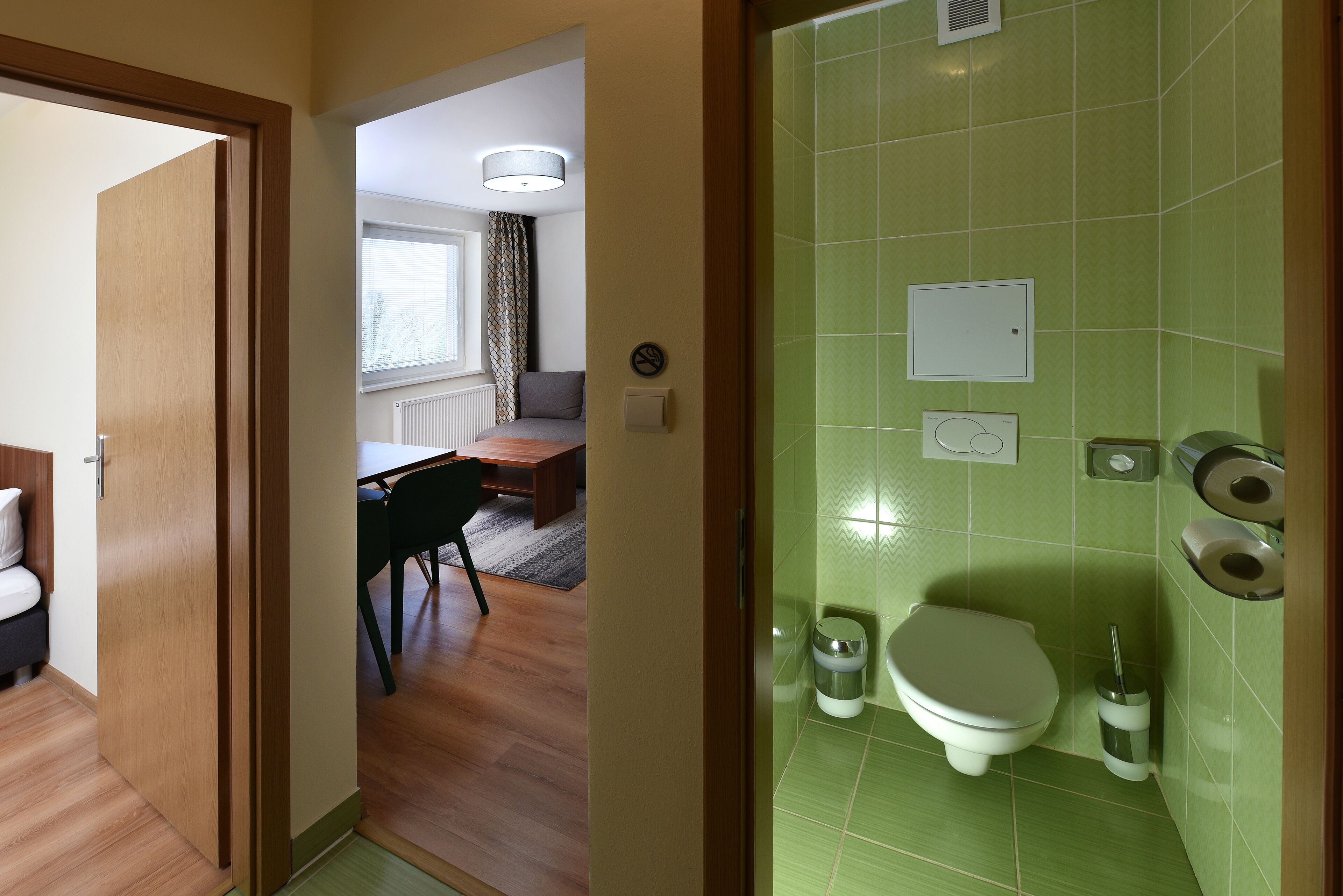 Habitación de hotel con puerta de madera, zona de comedor con mesa y sillas, y baño con azulejos verdes y un inodoro.