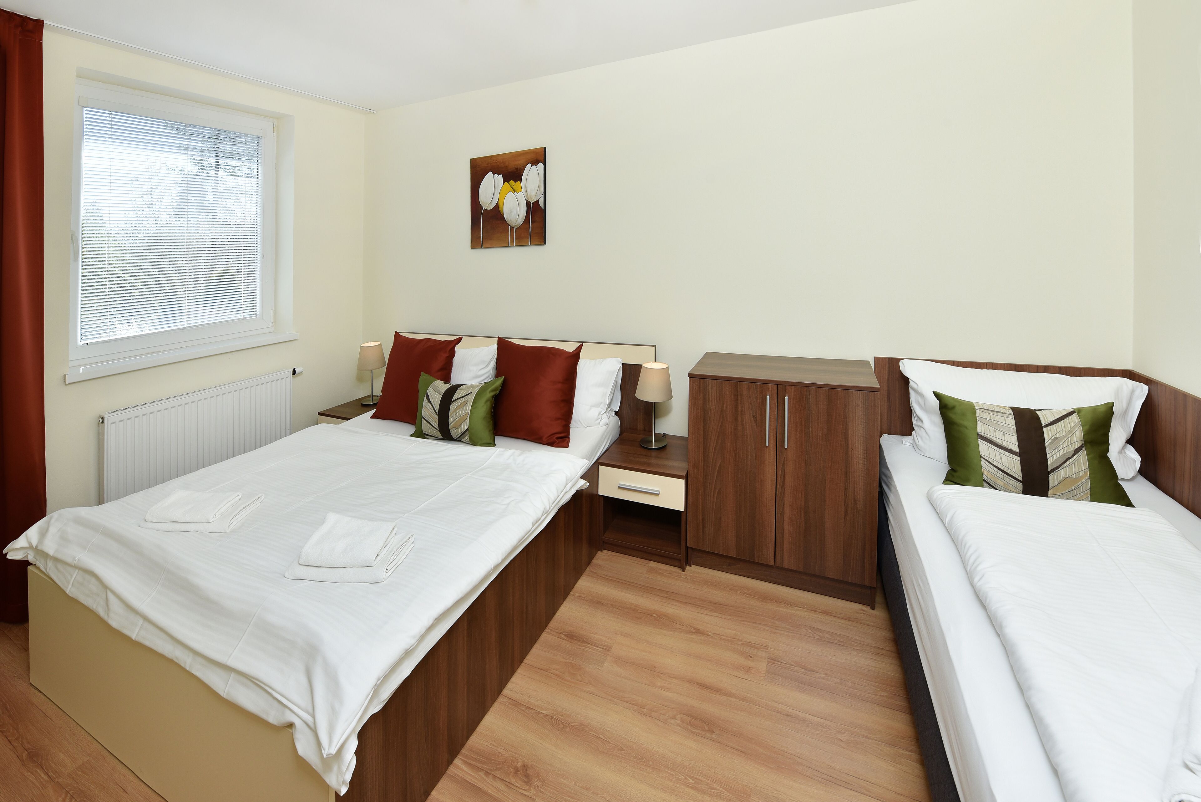 Une chambre d’hôtel avec deux lits, une armoire en bois, un radiateur, une fenêtre avec des stores et une œuvre d’art accrochée au mur.